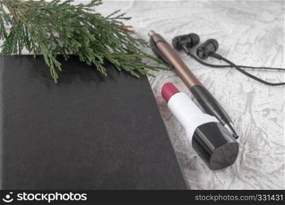 Green twig on a black notebook next to a pen, lipstick and headphones on a white background. There is a place for your text.. Green twig on a black notebook next to a pen, lipstick and headphones on a white background.
