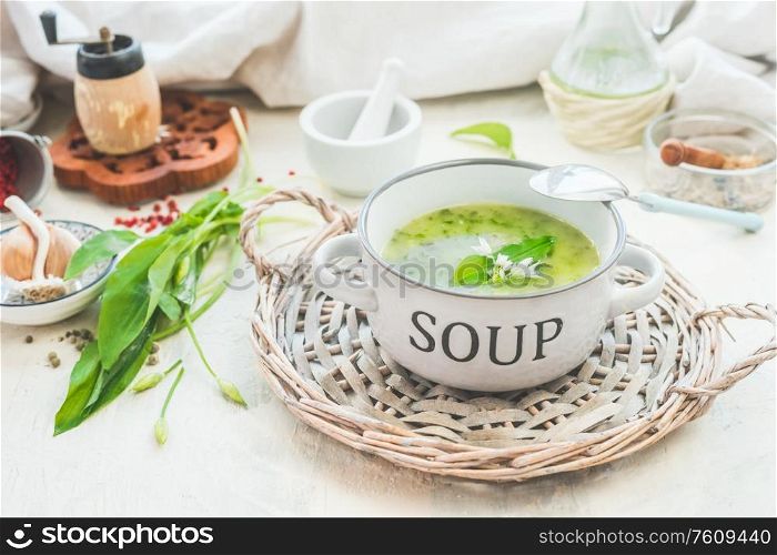 Green wild garlic soup in bowl with spoon on light table with ingredients. Close up. Seasonal home cuisine. Vegan food. Healthy cooking and eating