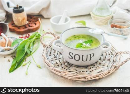 Green wild garlic soup in bowl with spoon on light table with ingredients. Close up. Seasonal home cuisine. Vegan food. Healthy cooking and eating