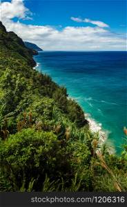 Green wild vegetation, blue water, abrupt shore line and beautiful clouds in the sky in this scene from Kalalau Trail, US. Stunning shore view from Kalalau Trail in Kauai, US
