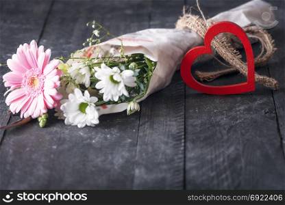 Greeting card design with a lovely bouquet of chrysanthemum flowers wrapped in a newspaper and a decorative wooden heart, displayed on a vintage black table.