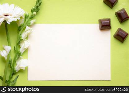 Greeting card design with lovely white flowers, pieces of chocolate and a blank message card, on a green background. A concept for celebrations and congratulations.