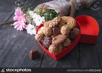 Greeting card idea with a heart-shaped box full of tasty cookies, chocolates and a bouquet of flowers wrapped in newspaper in the background, on a vintage table.