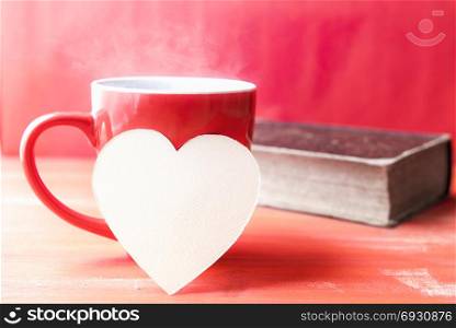 Greeting card idea with a red cup of hot drink with steam, a blank heart-shaped paper note leaned against it and an old book in the background, all in a red tone.