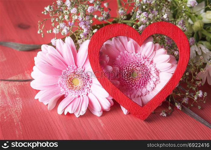 Greeting card idea with pink chrysanthemum and a red decorative heart leaned against them, on a red background. A concept for love, relationship, feelings, celebrations.