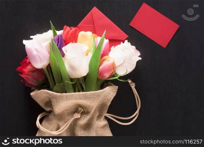 Greeting card with a bouquet of roses and tulips, wrapped in a hessian bag, a closed envelope and a red paper note, on a black wooden background.