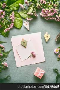 Greeting preparation with blank paper card mock up with heart and gift box on florist desktop with green leaves and flowers , top view, flat lay