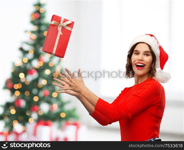 greetings and winter holidays concept - happy smiling young woman in santa helper hat catching red gift box over christmas tree lights background. happy young woman in santa hat catching gift box