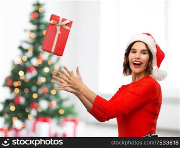 greetings and winter holidays concept - happy smiling young woman in santa helper hat catching red gift box over christmas tree lights background. happy young woman in santa hat catching gift box