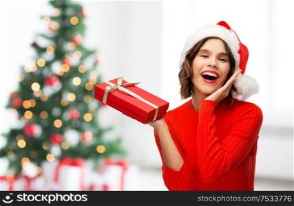 greetings and winter holidays concept - happy smiling young woman in santa helper hat with red gift box over christmas tree lights background. happy young woman in santa hat with red gift box