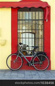 Grey bike in front of yellow facade and red window. Bicycle with trunk. Reflection of old building in the window. Window with grille.