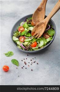 Grey bowl in motion with healthy fresh vegetables salad with lettuce and tomatoes, red onion and spinach on light table background with spatula spoon and fork