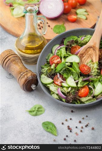 Grey bowl of healthy fresh vegetarian vegetables salad with tomatoes and cucumber, lettuce and spinach on light background with olive oil, mill and fresh vegetables on chopping board.