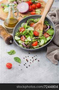 Grey bowl of healthy fresh vegetarian vegetables salad with tomatoes and cucumber, lettuce and spinach on light background with olive oil, mill and fresh vegetables on chopping board.