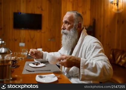 Grey-haired bearded senior man drinking tea during rest in sauna after health procedure. Senior man drinking tea rest in sauna