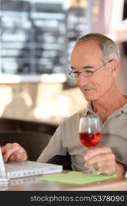 Grey haired man using laptop whilst drinking glass of wine