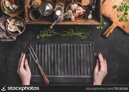 Grill or BBQ food background. Woman hold empty cast iron grill griddle and meat fork on roast rustic kitchen table. Wooden box with seasonings, oils and other kitchen utensils.Top view. Place for text