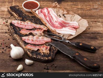 Grilled bacon strips on vintage wooden board with raw fresh smoked pork bacon on butchers paper with fork and knife on wood board with garlic and sauce.