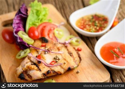 Grilled chicken on a Wood cutting board with salad, tomatoes, chilies cut into pieces, and sauce. Selective focus.