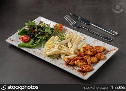 Grilled cubed chicken served with pasta and salad on white plate on dark stone table. chicken with pasta