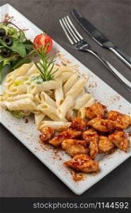 Grilled cubed chicken served with pasta and salad on white plate on dark stone table. chicken with pasta