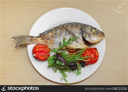 Grilled dorado fish with tomatoes, arugula and basil on white plate and wooden table&#xA;