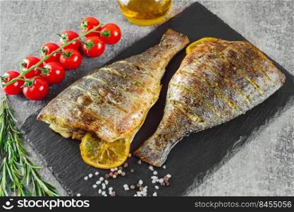 Grilled fish with roasted with lemon, rosemary, tomatoes, olive oil and spices on black slate dish, on grey background.. Grilled fish with roasted with lemon and spices
