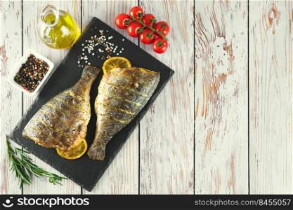 Grilled fish with roasted with lemon, rosemary, tomatoes, olive oil and spices on black slate dish, on white wooden background. Top view, flat lay with copy space.. Grilled fish with roasted with lemon and spices