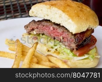 Grilled hamburger with bacon, shredded lettuce, tomatoe slice. Meat on bread roll with fried potatoes on outdoor, wrought iron patio table.