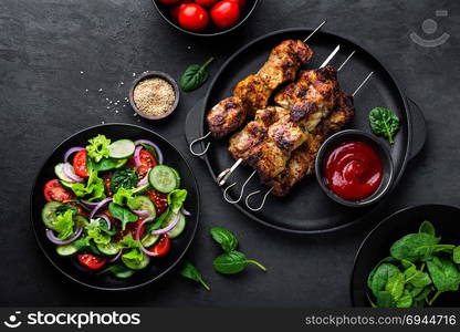 Grilled meat skewers, shish kebab and healthy vegetable salad of fresh tomato, cucumber, onion, spinach, lettuce and sesame on black background, top view