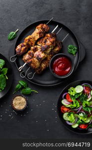 Grilled meat skewers, shish kebab and healthy vegetable salad of fresh tomato, cucumber, onion, spinach, lettuce and sesame on black background, top view