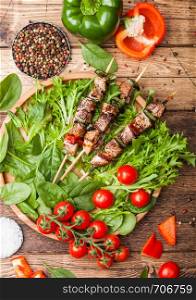 Grilled pork and chicken kebab with paprika in round wooden plate of lettuce salad, on wooden background with tomatoes and spinach.