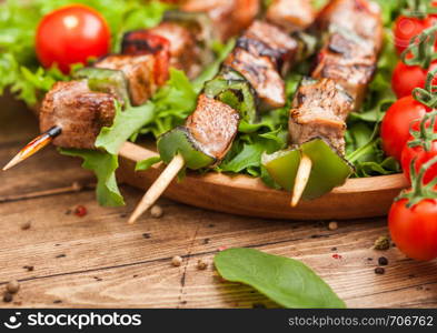 Grilled pork and chicken kebab with paprika in round wooden plate of lettuce salad, on wooden background with tomatoes and spinach.