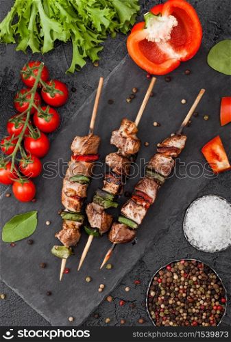 Grilled pork and chicken kebab with paprika on stone chopping board with salt, pepper and tomatoes on black background. Fresh lettuce and paprika pepper.