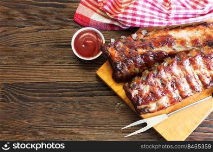 grilled pork ribs on a wooden cutting board with kitchen fork for meat and tomato ketchup on brown wooden surface. Top view, flat lay.. grilled pork ribs on a cutting board