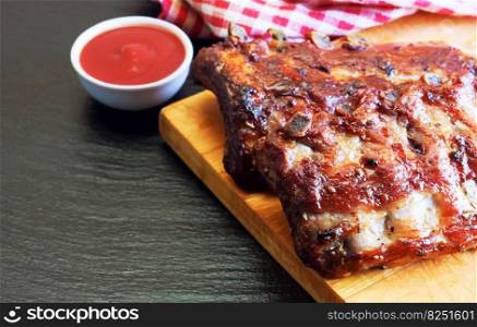 grilled pork ribs on a wooden cutting board with tomato ketchup on black slate surface. grilled pork ribs on a cutting board