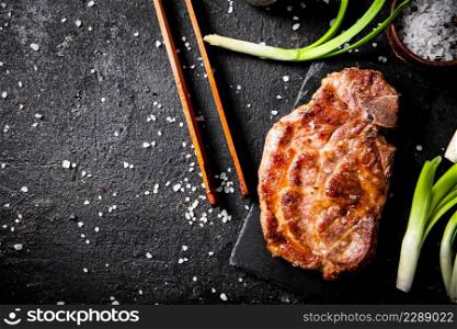 Grilled pork steak on a stone board with green onions and spices. On a black background. High quality photo. Grilled pork steak on a stone board with green onions and spices.