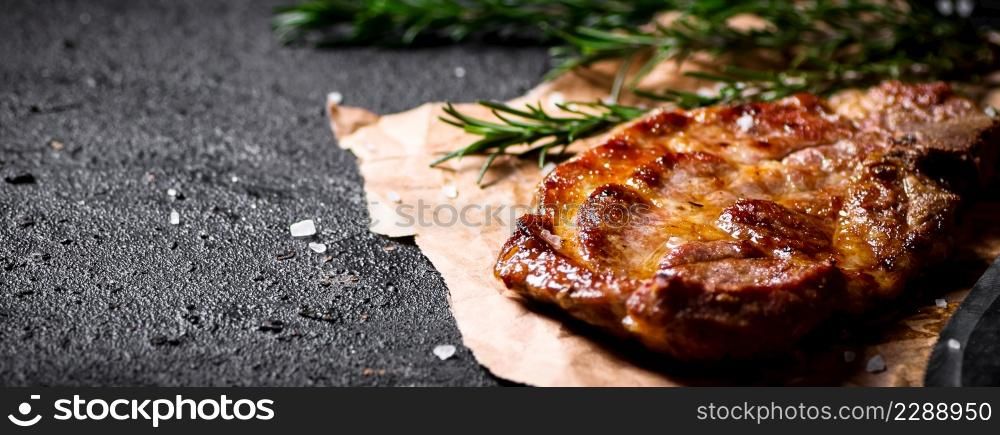 Grilled pork steak on paper with rosemary. On a black background. High quality photo. Grilled pork steak on paper with rosemary.