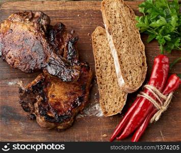 grilled pork steaks on the bone on a kitchen cutting board, baked sliced rye flour bread and red chili pepper, top view