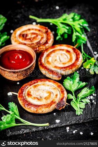 Grilled sausages on a stone board with tomato sauce and parsley. On a black background. High quality photo. Grilled sausages on a stone board with tomato sauce and parsley.