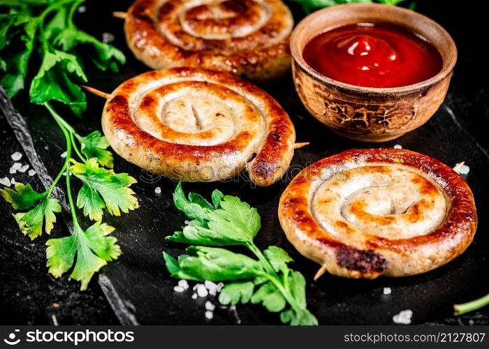 Grilled sausages on a stone board with tomato sauce and parsley. On a black background. High quality photo. Grilled sausages on a stone board with tomato sauce and parsley.