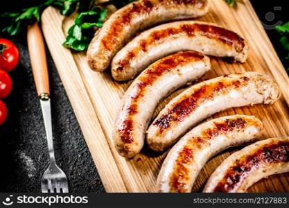 Grilled sausages on a wooden cutting board with parsley and tomatoes. On a black background. High quality photo. Grilled sausages on a wooden cutting board with parsley and tomatoes.