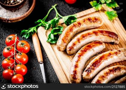 Grilled sausages on a wooden cutting board with parsley and tomatoes. On a black background. High quality photo. Grilled sausages on a wooden cutting board with parsley and tomatoes.