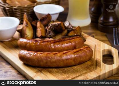 Grilled sausages with roast potato with spices on wooden board with light beer on table