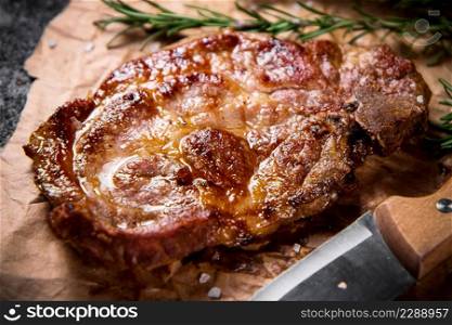 Grilled steak on paper with a knife and a sprig of rosemary. Against a dark background. High quality photo. Grilled steak on paper with a knife and a sprig of rosemary.
