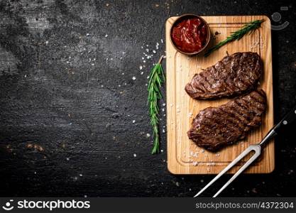 Grilled steak with tomato sauce and rosemary on a cutting board. On a black background. High quality photo. Grilled steak with tomato sauce and rosemary on a cutting board.
