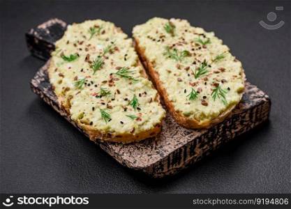 Grilled toast with avocado paste, eggs, cream cheese, salt, spices and herbs on a textured concrete table