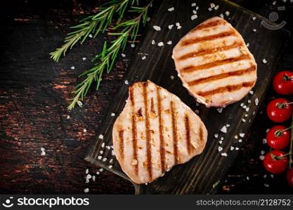 Grilled tuna steak on a cutting board with spices, tomatoes and rosemary. Against a dark background. High quality photo. Grilled tuna steak on a cutting board with spices, tomatoes and rosemary.