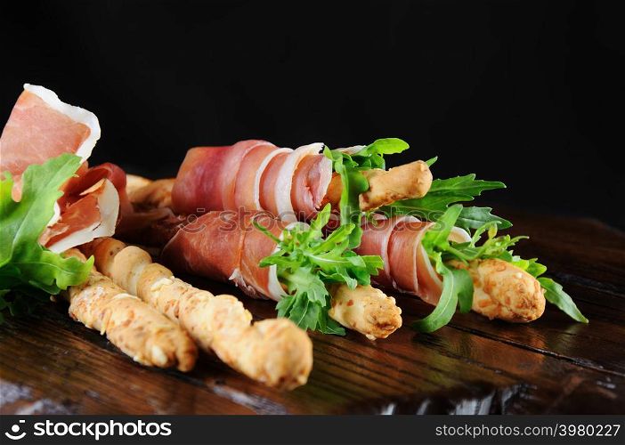 Grissini - bread sticks with Parmesan, wrapped with a piece of prosciutto and arugula. Italian dish with antipast on a wooden table.