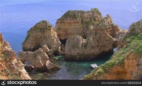 Grnn bewachsene Felseninsel im flachen tnrkisblauen Meer mit Steinen / zwei Boote ankern in der kleinen Bucht; Knste der Algarve, Portugal.